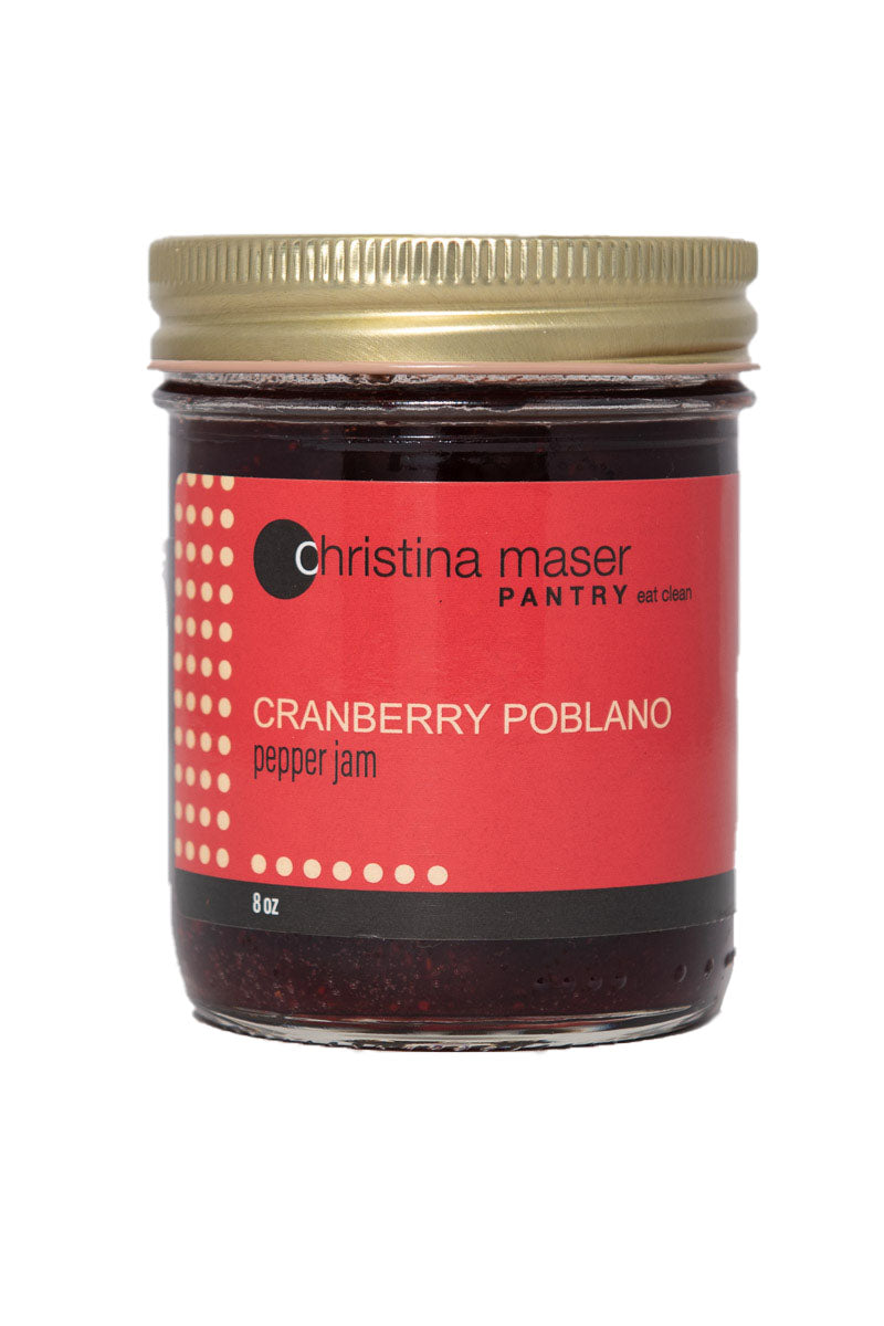 Cranberry Poblano Pepper jam. Full of sweet heat. A clear glass jar with a red wraparound label with beige accents.